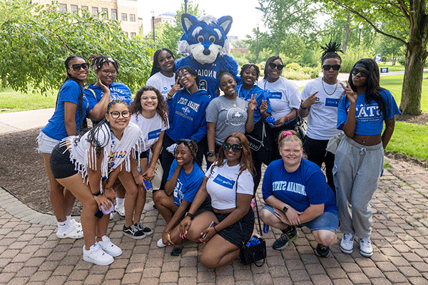 A diverse group of 14 smiling female students pose in front of Sycamore Sam on a campus walkway at Indiana State. Everyone is wearing blue or white shirts. Those in the back row stand beside Sam while those in the front row kneel in front.